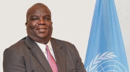 A man in a brown suit standing in front of the blue UN flag