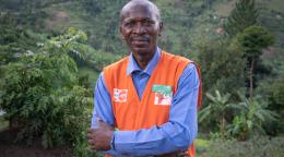 A man in a blue shirt and orange vest stands with his arms crossed in front of a green leafy background.