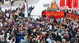 A crowd of protestors, carrying signs gather in front of a platform where people hold up a red poster demanding the end of fossil fuels