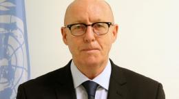 Man in dark suit stands in front of UN flag against white wall looking into the camera
