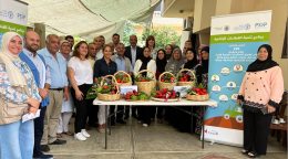 A group of people, men and women, in different coloured clothes pose for a picture behind a table where various fruits and vegetables are displayed.