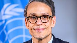 A man in a blue suit and black spectacles stands in front of a blue UN flag