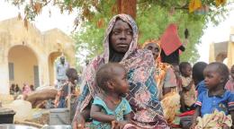 一位非洲妇女抱着孩子坐在乍得一所中学的院子里，身后都是其他妇女与儿童