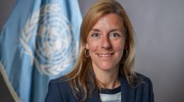 A woman with light brown hair wearing a dark blue suit stands against a grey background with a blue UN flag.
