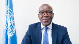 A man in a dark navy suit and blue tie with black spectacle frames sits in front of a blue flag with a white UN logo.