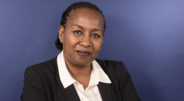 A headshot of a woman in a black suit and a white shirt against a blue background