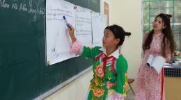 A girl in a colourful green dress points to a white board in a classroom while another woman in a pink dress, likely a teacher, watches on.