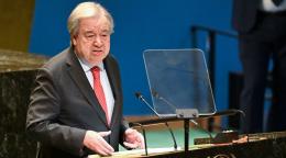 A man in a black suit and red tie stands at a podium and speaks into a mic at the UN General Assembly