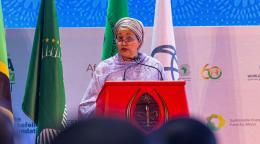 A woman in a purple dress and headscarf, the UN Deputy Secretary-General, Amina Mohammed speaks at a podium against a colourful background