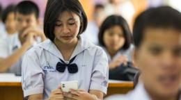 A girl student in a white school uniform checks her mobile phone while sitting in a classroom