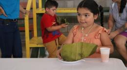 A little girl in an orange shirt sits at a table with a plate of tamales wrapped in leaves and a cup of liquid next to it. Behind her in a boy in a red shirt.