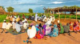 A panoramic view of a group of community members gathered. The women sit across from the men, wearing colourful full length clothes, wile the men sit on the other side dressed in white and with head-gears.