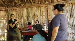 A woman in a black shirt speaks to two other women in a thatched house