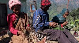 A woman and two men are sitting together weaving bamboo strands.