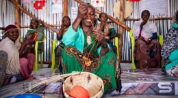A group of women in colorful traditional clothing are gathered in a circle weaving.
