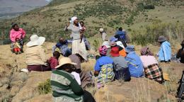 A woman stands in the middle of a group outdoors and speaks to them, while holding her child
