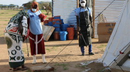 两名非洲妇女和一名非洲男性站在黄土地上，身旁是简易的房屋。