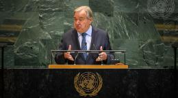 A man, the Secretary-General of the United Nations, in a dark suit and tie, gesticulates as he addresses people from a podium