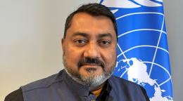 A man in a dark suit stands in front of a blue flag with the UN logo on it.
