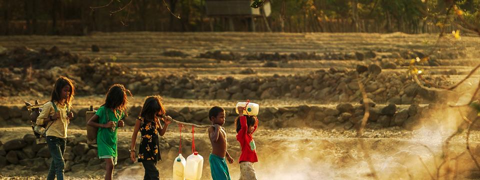 Little children walk in a line as they all hold a large stick holding jugs of water. 