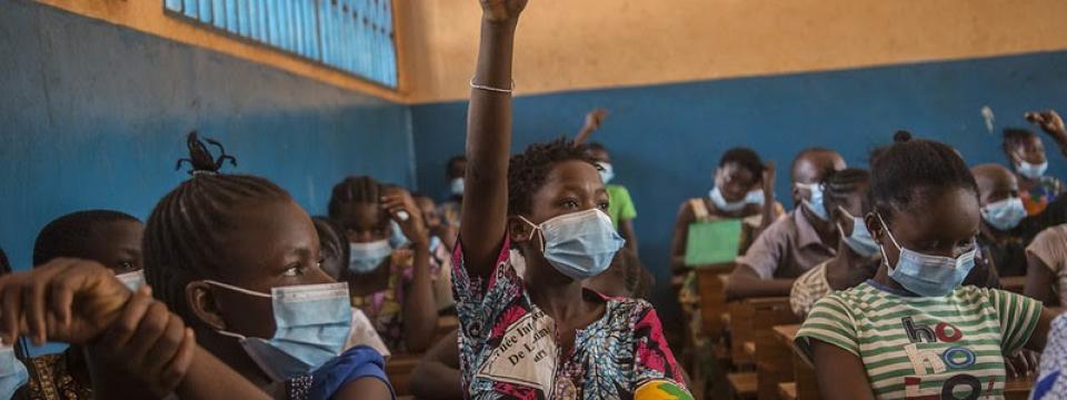 In a classroom of students wearing face masks, one student proudly raises their hands.