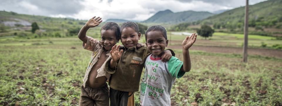 Three smiling boys in a field wave at the camera. 