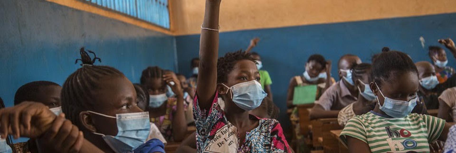 In a classroom of students wearing face masks, one student proudly raises their hands.