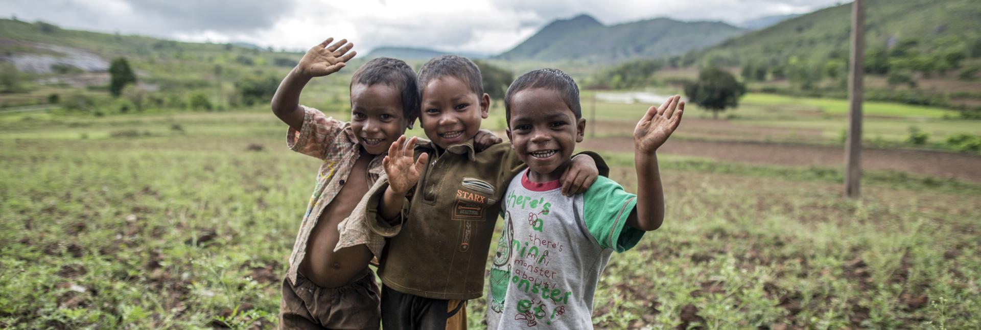 Three smiling boys in a field wave at the camera. 
