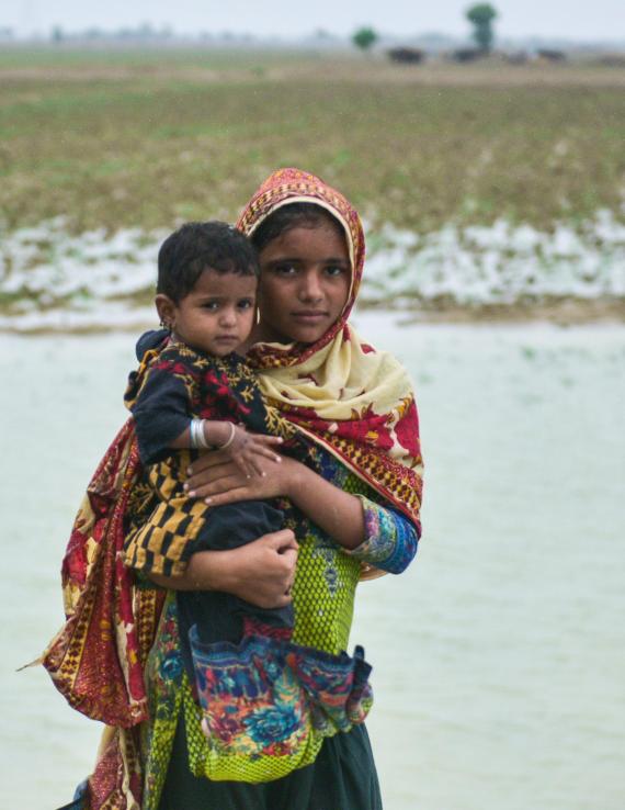 A girl in scarf holds a toddler in her arms in front of a pool of water collected on a field, against a grey sky. 