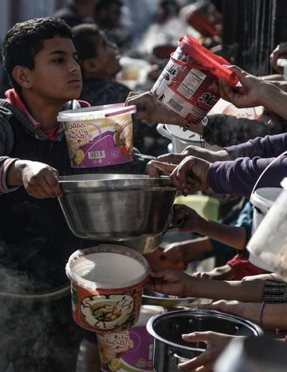 A young boy hands a large metal bowl, presumably with food, to people, including youth, lined up for food and water.