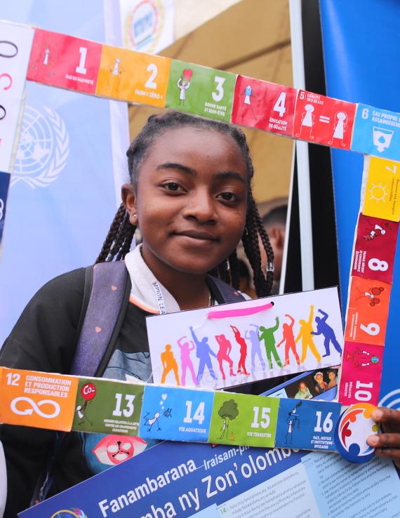 A young girl in a school uniform holds a frame with the SDGs, she looks at the camera and smiles