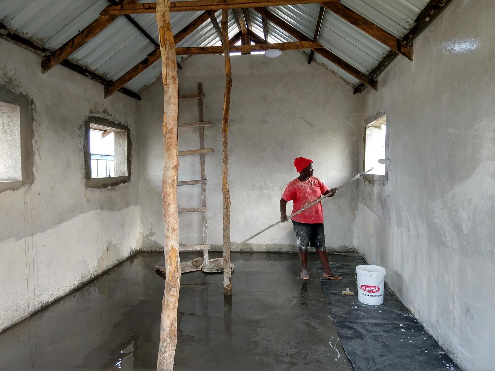 Человек в красной рубашке и кепке красит стены здания. 