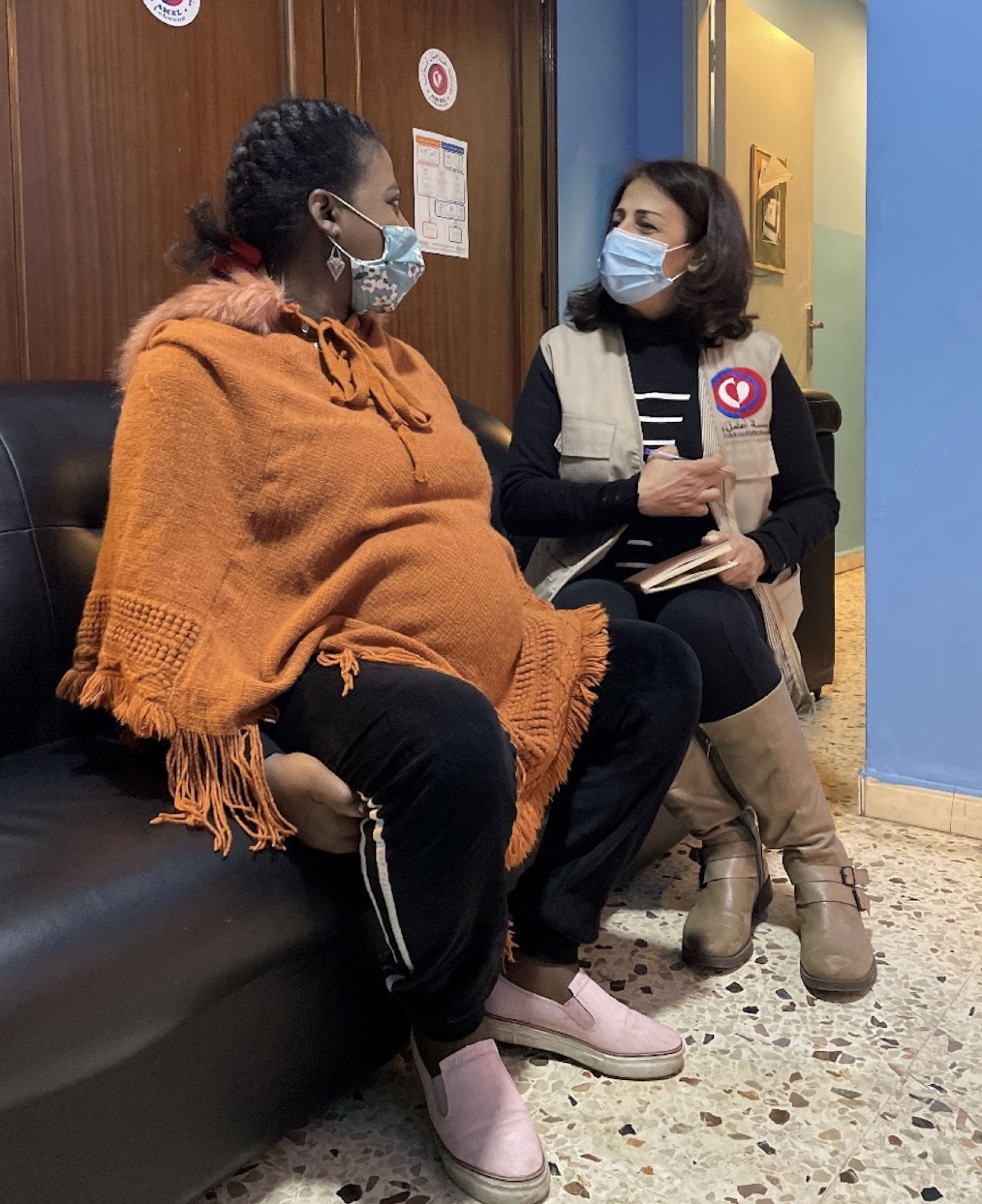 Two women, one in an orange sweater and one in a black shirt and tan vest, sit on a couch talking to each other. 