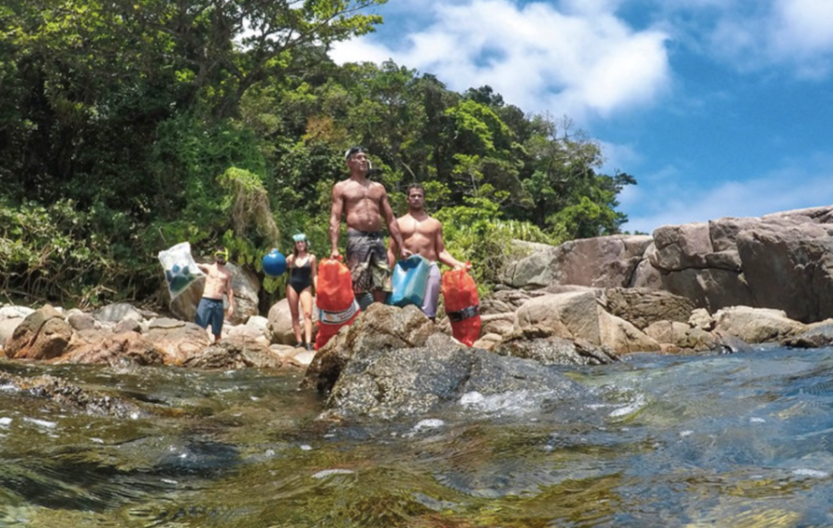 Several people, with orange and white bags, stand on rocks near the ocean. 