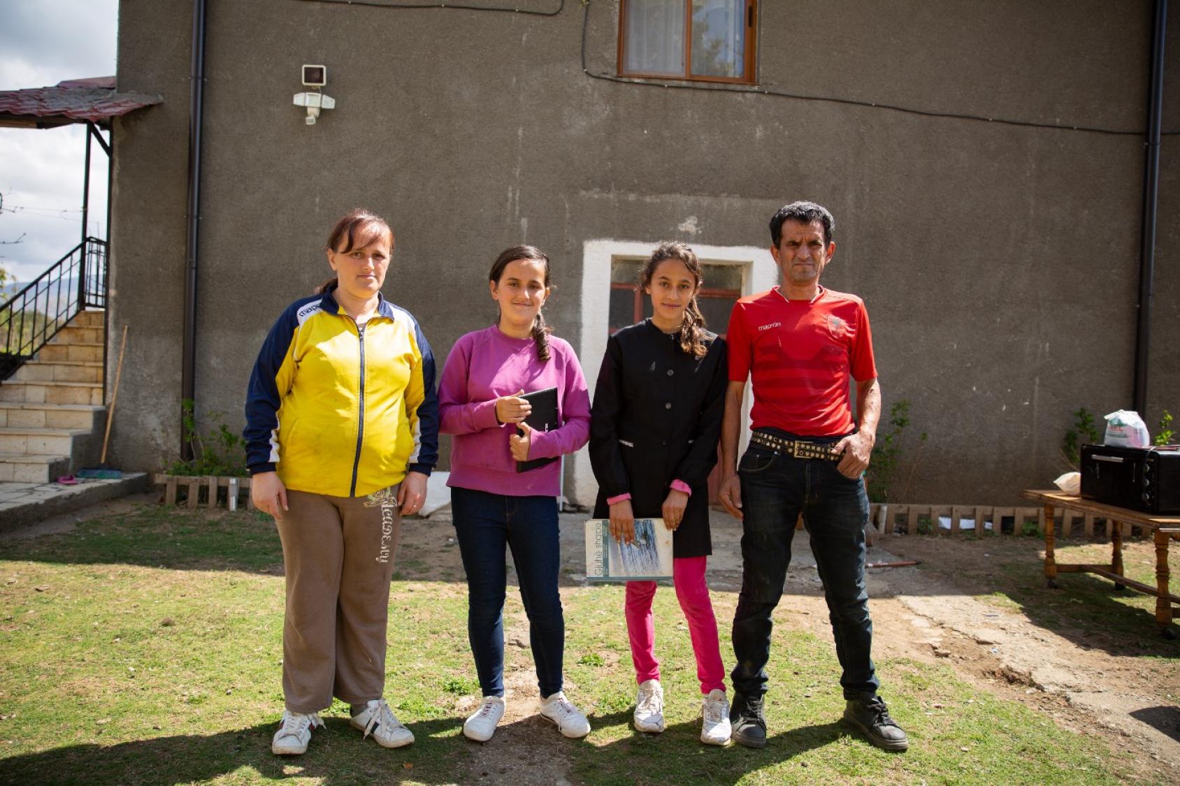 A family of four, including a man, a woman, and two daughters, stand together in front of a building.