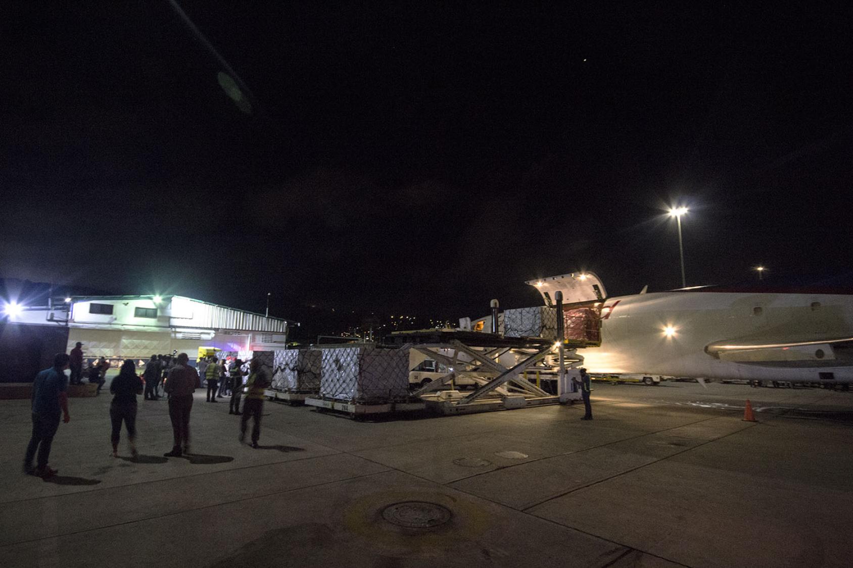 Toma nocturna de un área de aterrizaje, donde se aprecia a un grupo de personas y un cargamento de vacunas facilitadas por el mecanismo COVAX, en el lateral de un avión.