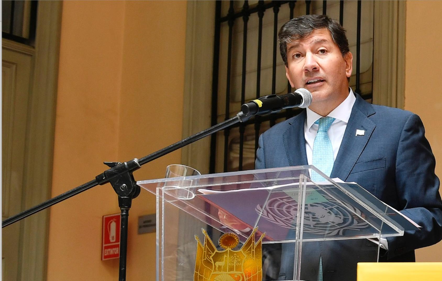 Un homme vêtu d'un costume bleu et d'une cravate bleu clair s'exprime au microphone devant un public que l'on devine assis dans la salle derrière l'objectif.