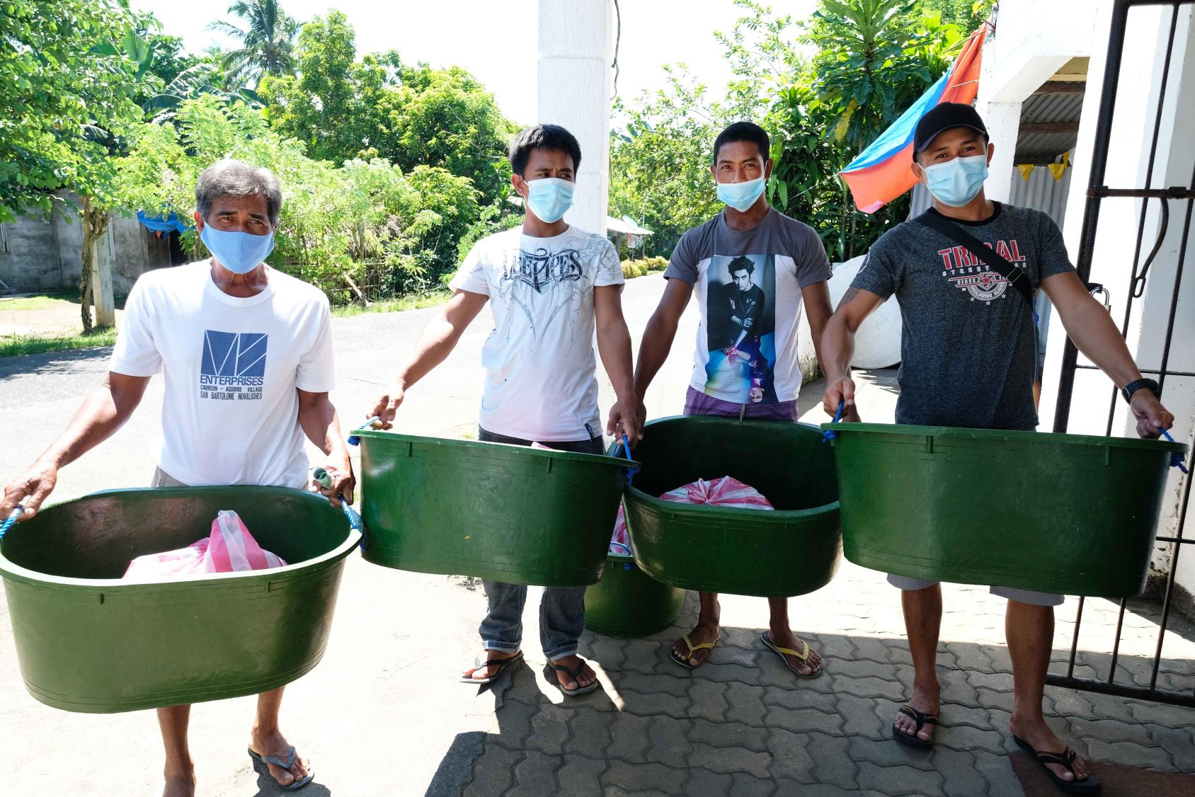 Cuatro hombres parados uno al lado del otro, cada uno carga un gran cubo de basura verde, que llevan entre las manos.
