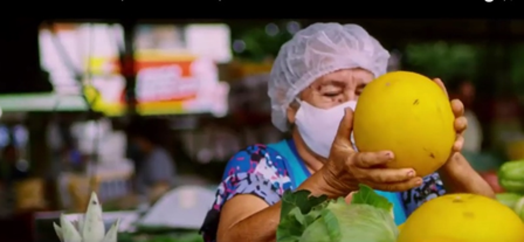 Une femme portant une charlotte et un masque de protection respiratoire blanc dépose un gros fruit jaune sur son étal de fruits et légumes.