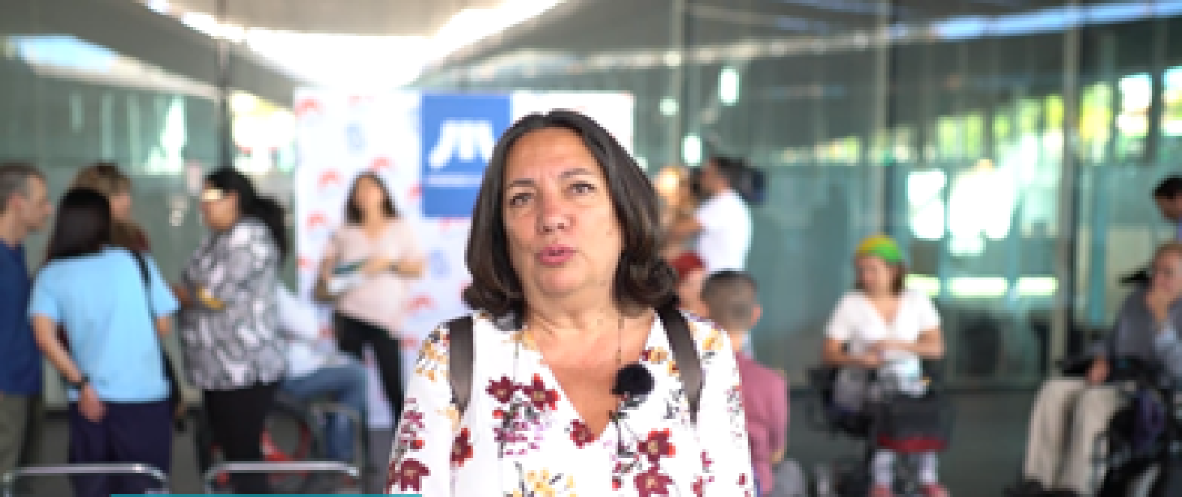 Une femme portant une chemise à fleurs s'exprime face à la caméra. Elle se tient debout dans le hall d'une station de métro.