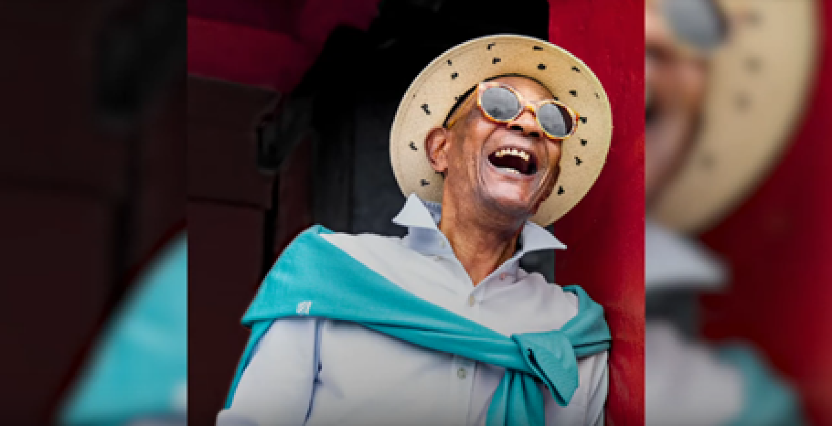 Photographie d'un homme portant une chemise bleu ciel, un gilet bleu autour des épaules, un chapeau de paille et des lunettes de soleil et éclatant de rire.