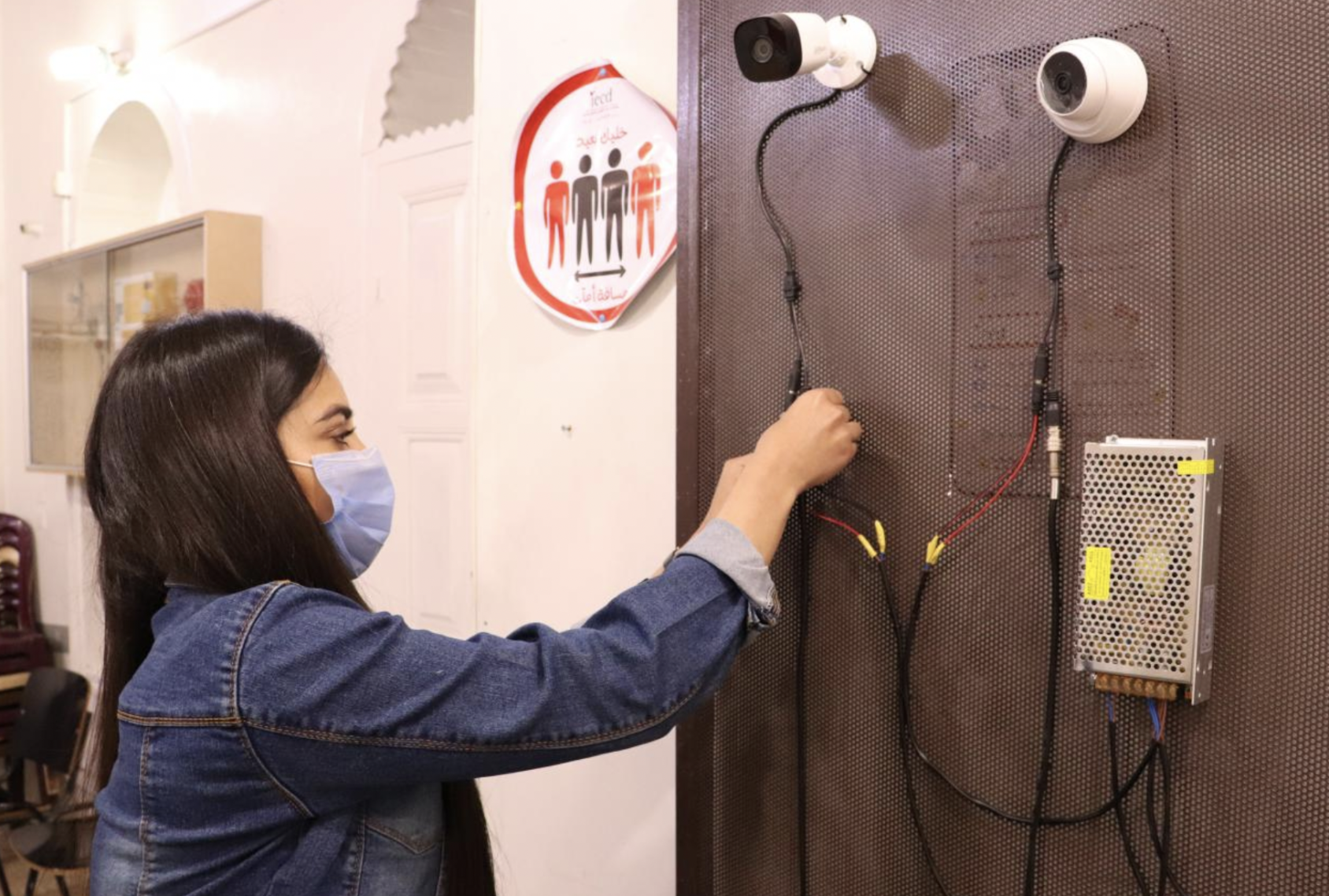 Una chica con una mascarilla azul conecta los cables de la cámara en una pared.
