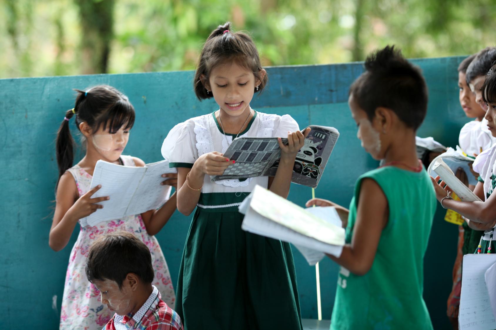 Un grupo de niños y niñas leen libros, de pie, en la sede de una escuela.