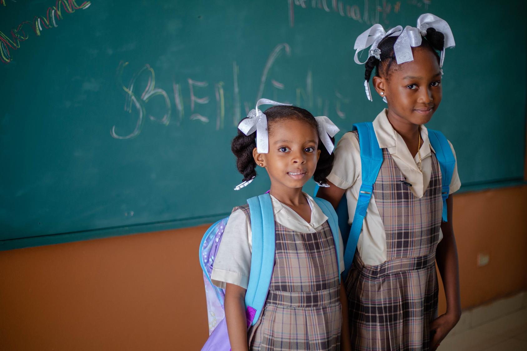 Two girls wearing backpacks and school uniforms stand in front of a chalkboard.