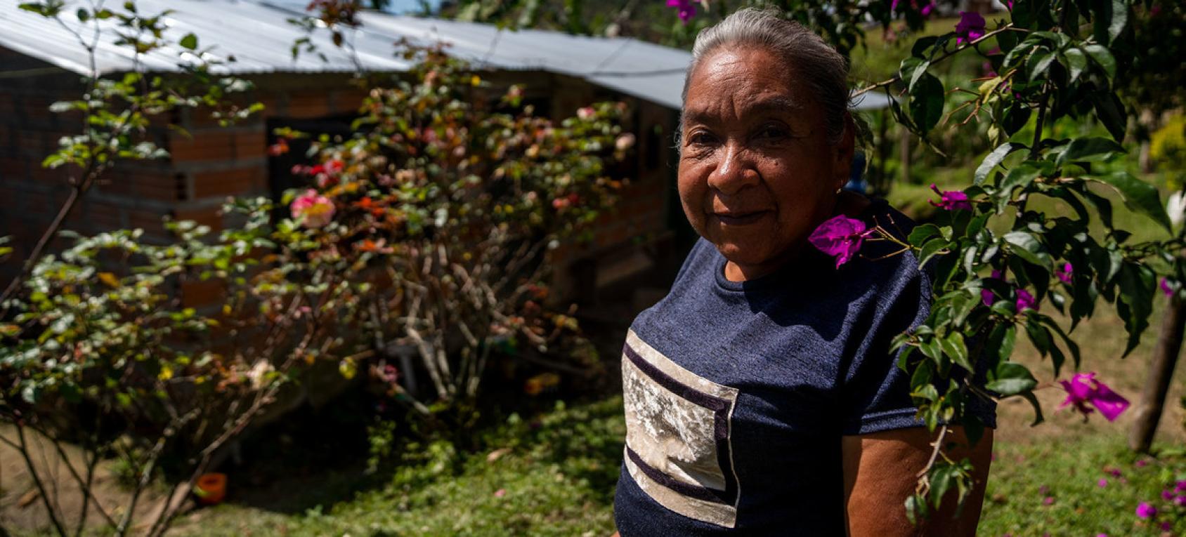 An older woman stares directly at the camera as she stands near flowering trees.