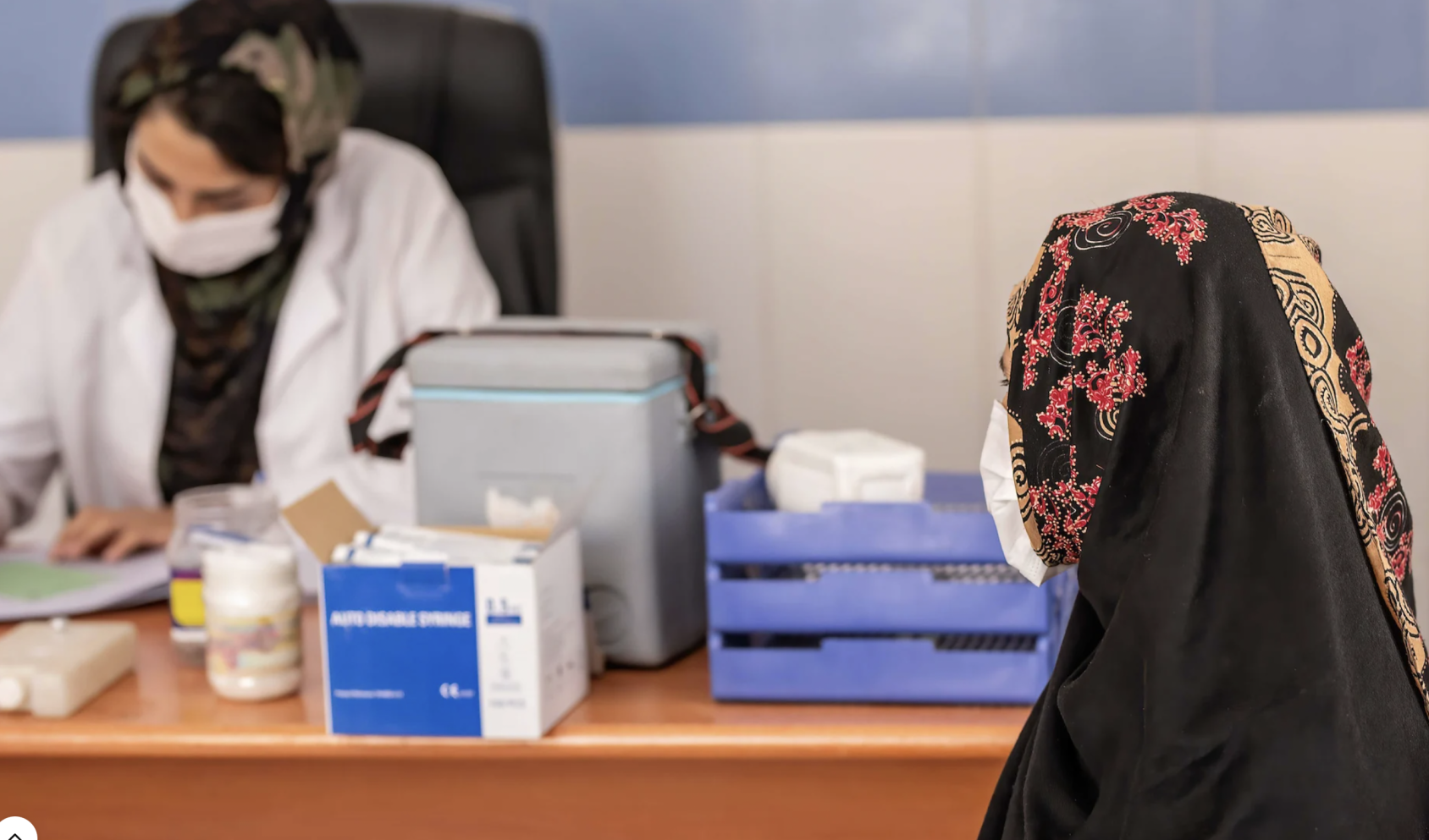 A woman sits near a health care professional writing at her desk. 