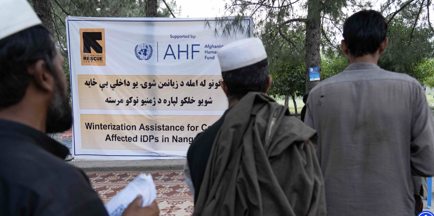 Three men stand in line near a big sign in Arabic and English. 