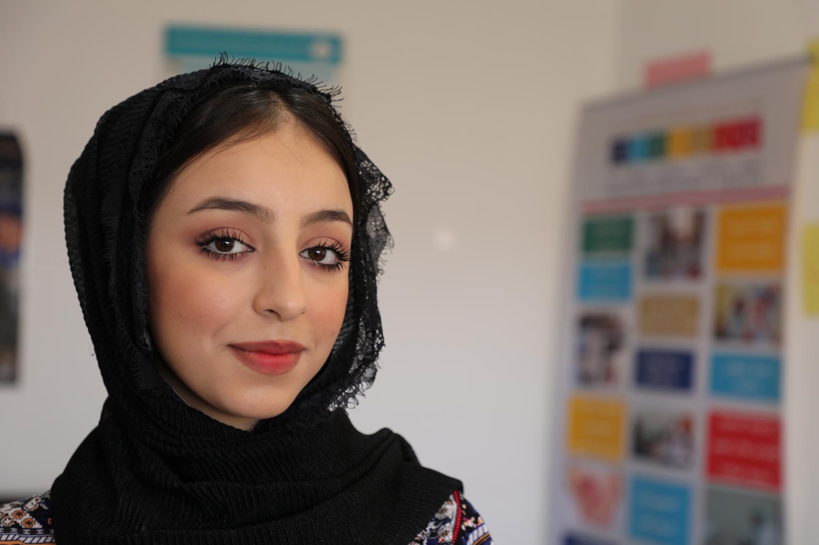 A woman looks directly at the camera with colorful cards on the wall, in the background.  