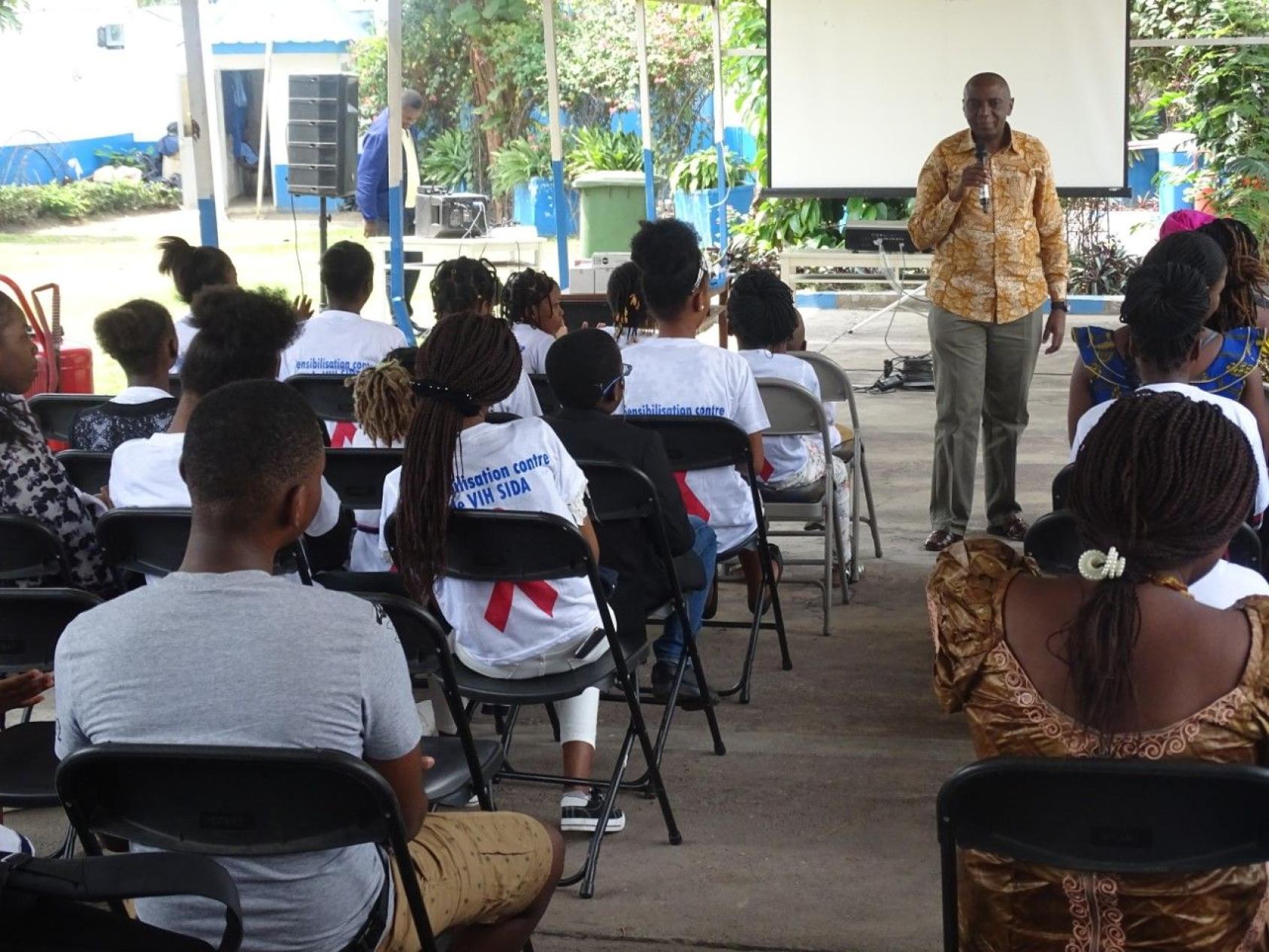 De jeunes femmes et hommes assis sur des chaises assistent à une session de sensibilisation au VIH/Sida tandis qu'un orateur, debout devant un tableau blanc, s'exprime à un micro.
