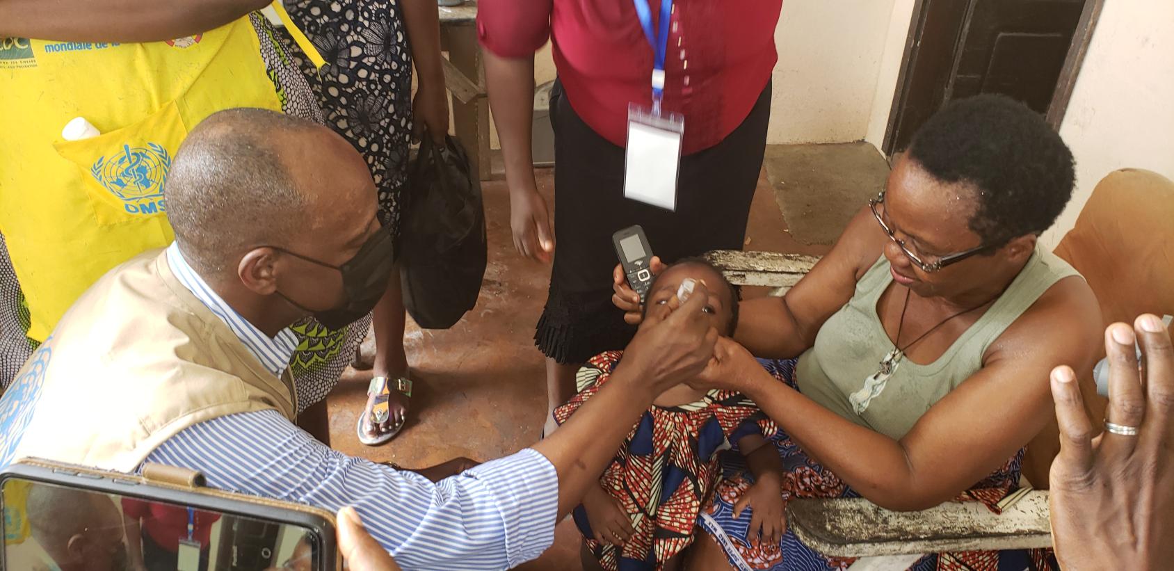 Un homme portant un masque de protection respiratoire administre le vaccin de la polio à un jeune enfant tenu par sa grand-mère.